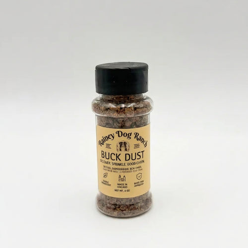 A bottle of Buck Dust dog topper by Rainey Dog Ranch, featuring a tan label and a black cap, set against a plain light gray background.